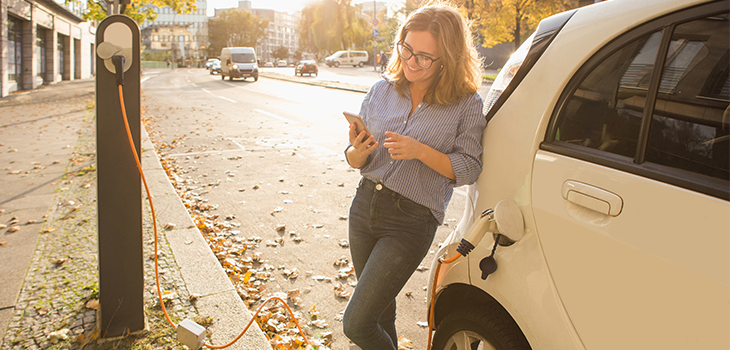 Young woman is standing near the electric car and looks at the smart phone. The rental car is charging at the charging station for electric vehicles.