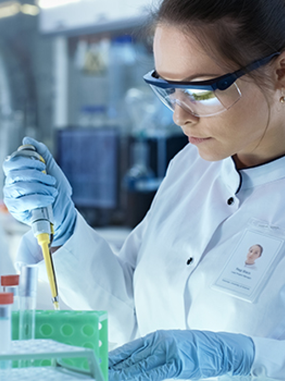 Personne en blouse blanche et gants bleus manipulant une micropipette dans un laboratoire, avec des tubes à essai dans un support vert, un microscope et divers équipements scientifiques en arrière-plan.