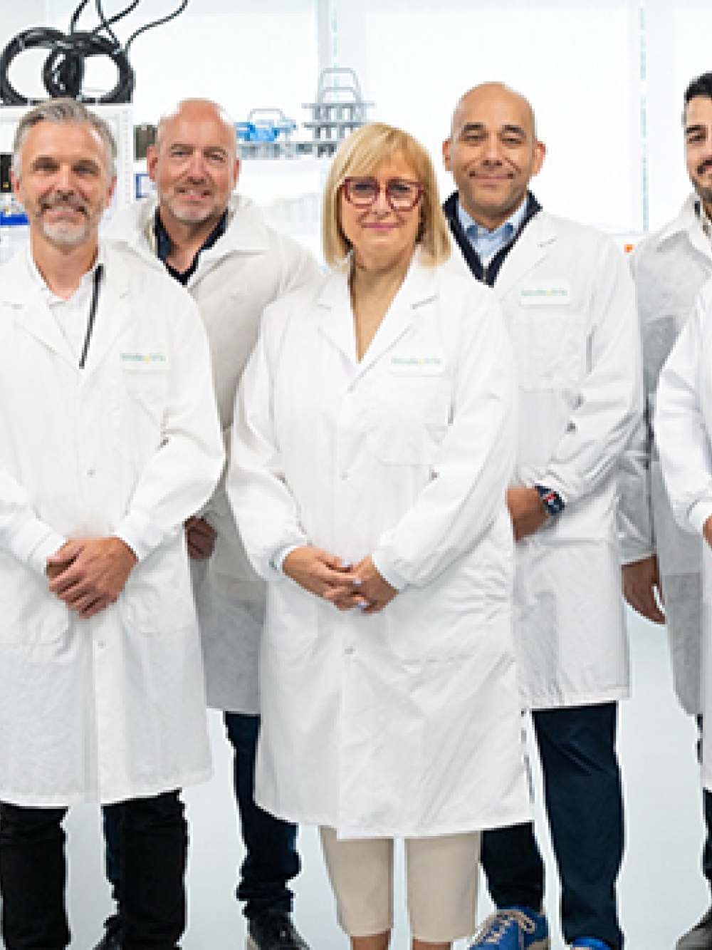 Groupe de personnes portant des sarraus blancs dans un laboratoire moderne, entouré d’équipements scientifiques, d’étagères avec produits et d’un grand réfrigérateur vitré.