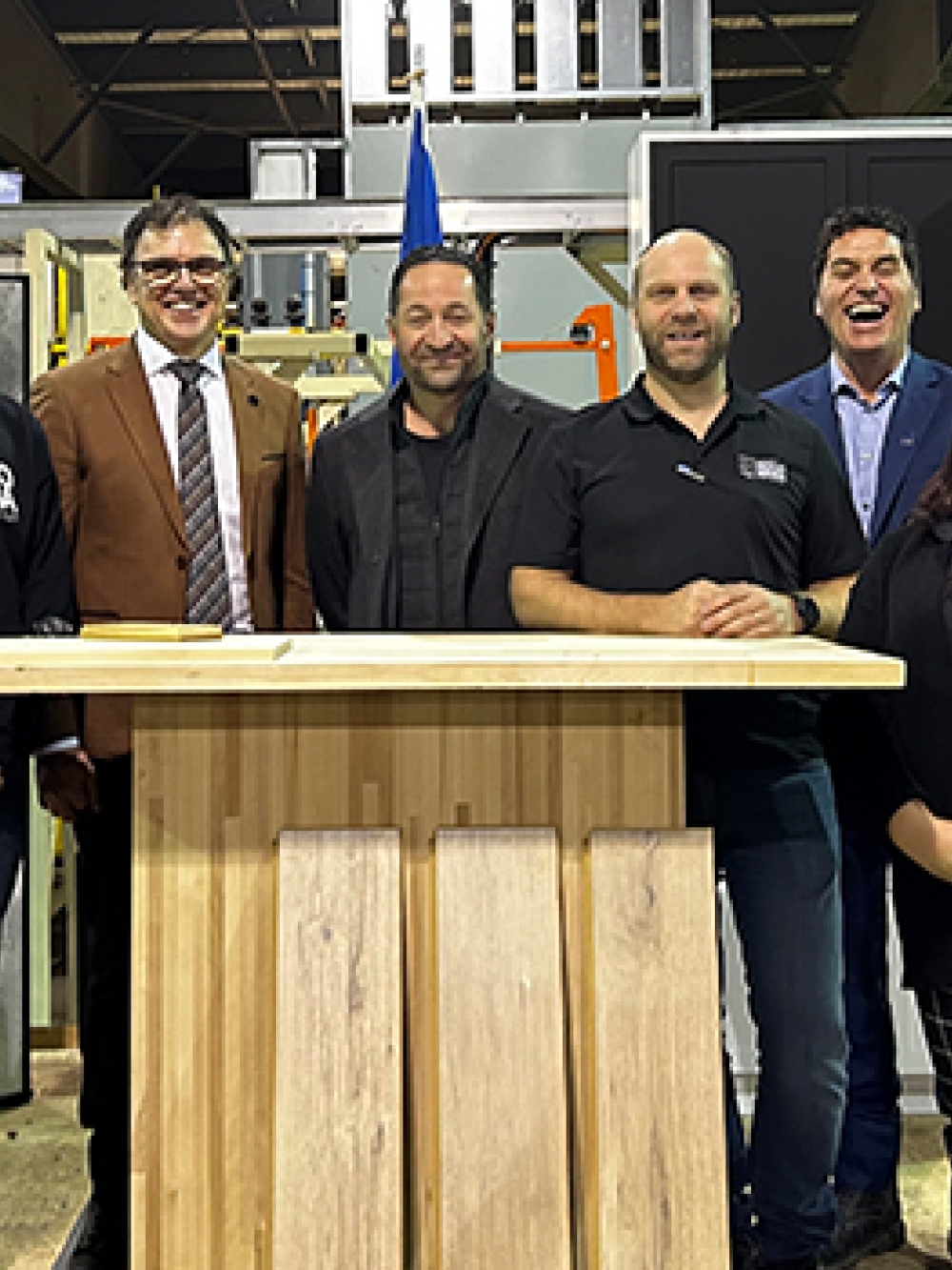 Groupe de personnes debout derrière un lutrin en bois dans un atelier industriel, avec des équipements et matériaux de scierie visibles en arrière‑plan ainsi qu’un écran de présentation sur la gauche.