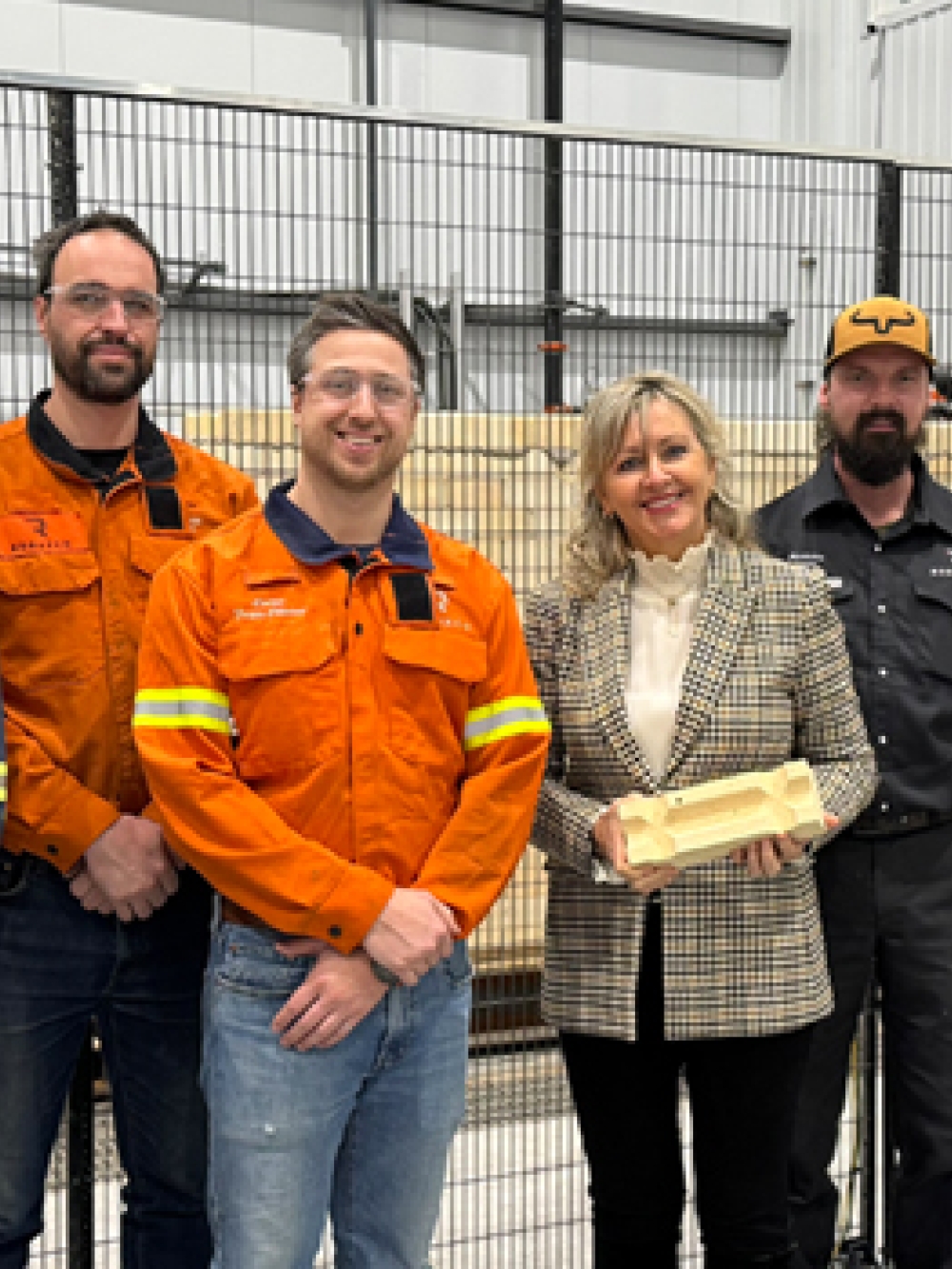 Groupe de neuf personnes posant dans une usine, dont plusieurs portent des vêtements de travail orange ou foncés, avec des équipements industriels et des robots derrière une grille de sécurité.