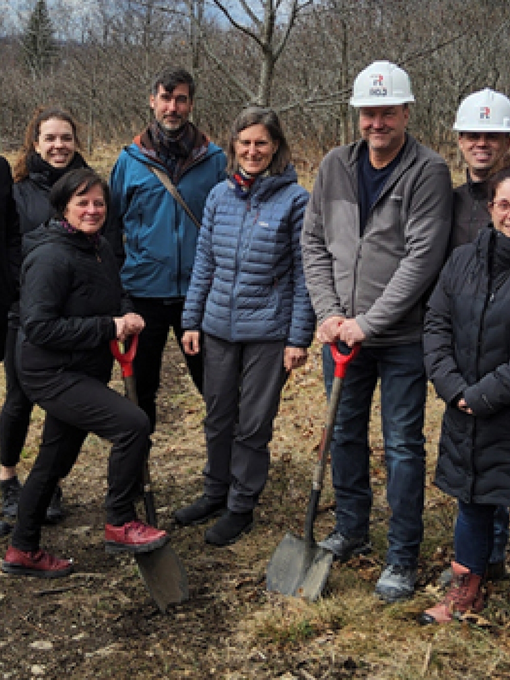 Groupe de personnes debout à l’extérieur dans un milieu naturel, tenant des pelles lors d’une pelletée de terre symbolique sur un terrain boisé.
