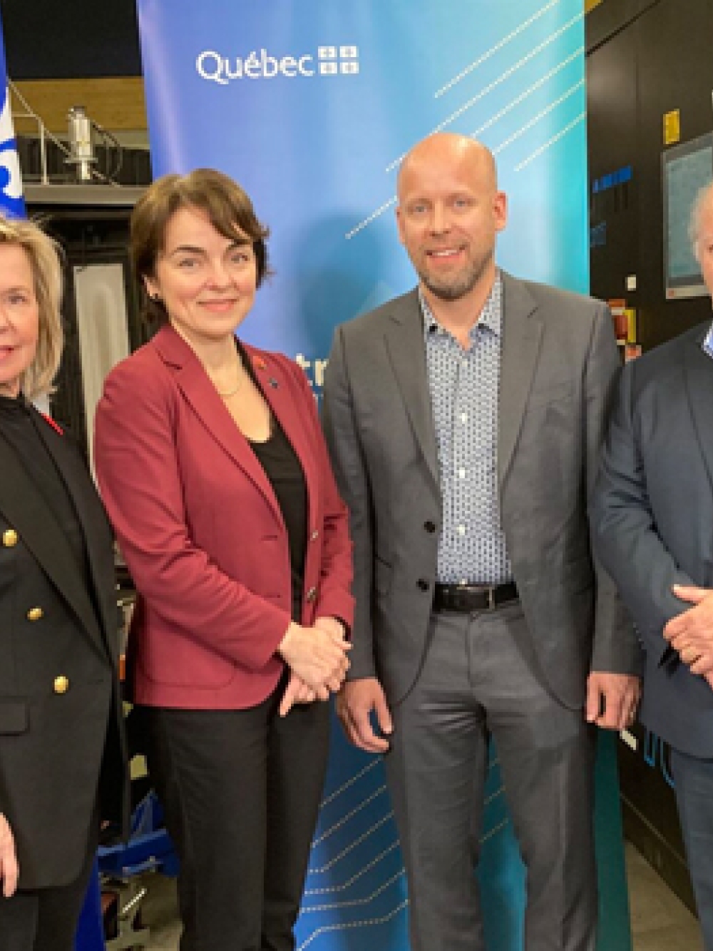 Groupe de personnes en tenue professionnelle posant dans un atelier industriel, devant un panneau bleu portant le logo du Québec et des équipements de machinerie en arrière-plan.