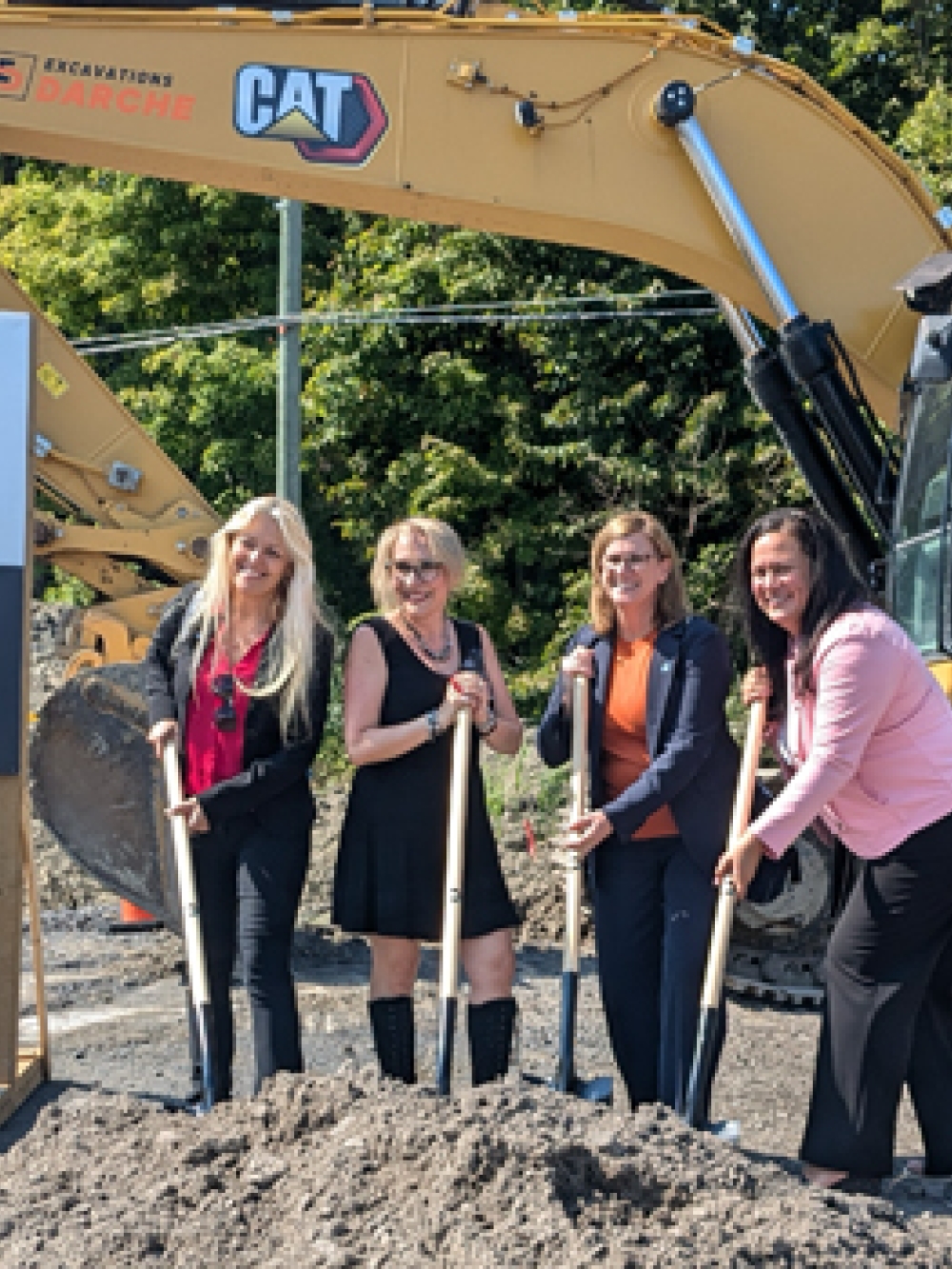 Quatre personnes tenant des pelles lors d’une cérémonie de première pelletée devant un panneau EcoloPharm et une pelle mécanique CAT sur un chantier.