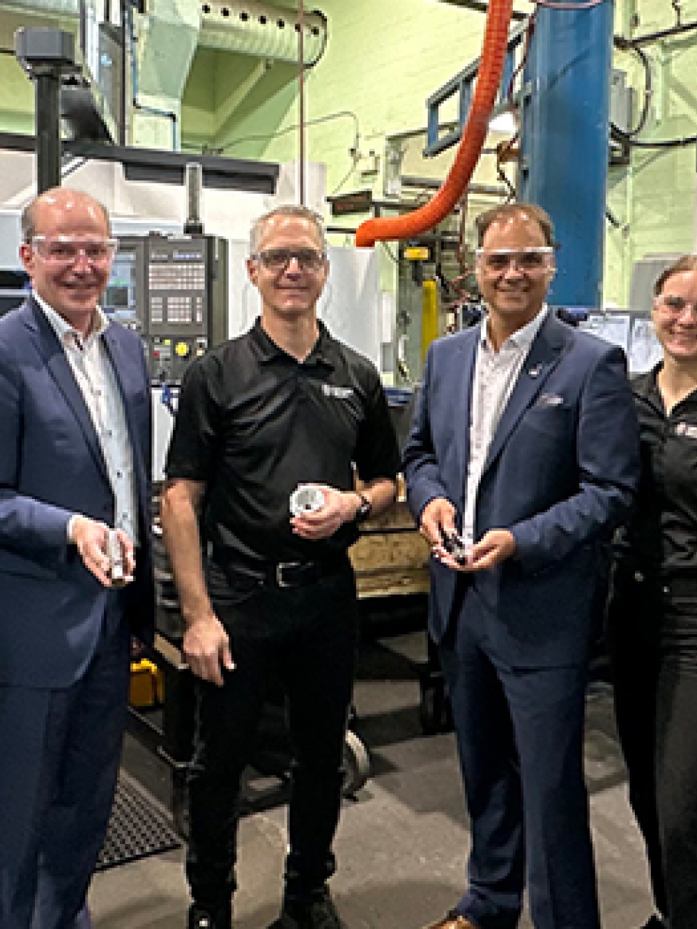 Groupe de personnes debout dans un atelier industriel, entourées de machines-outils et d’équipements de fabrication, certaines tenant des pièces mécaniques métalliques.