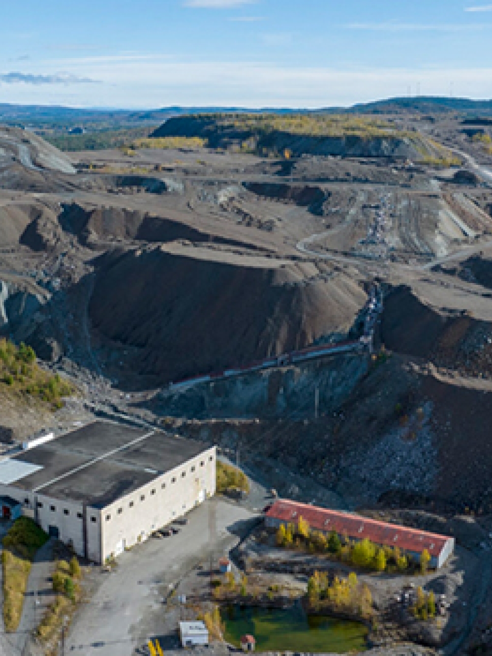 Vue aérienne d’un site minier avec des bâtiments industriels entourés de vastes amas de résidus et de chemins de gravier, dans un paysage vallonné.