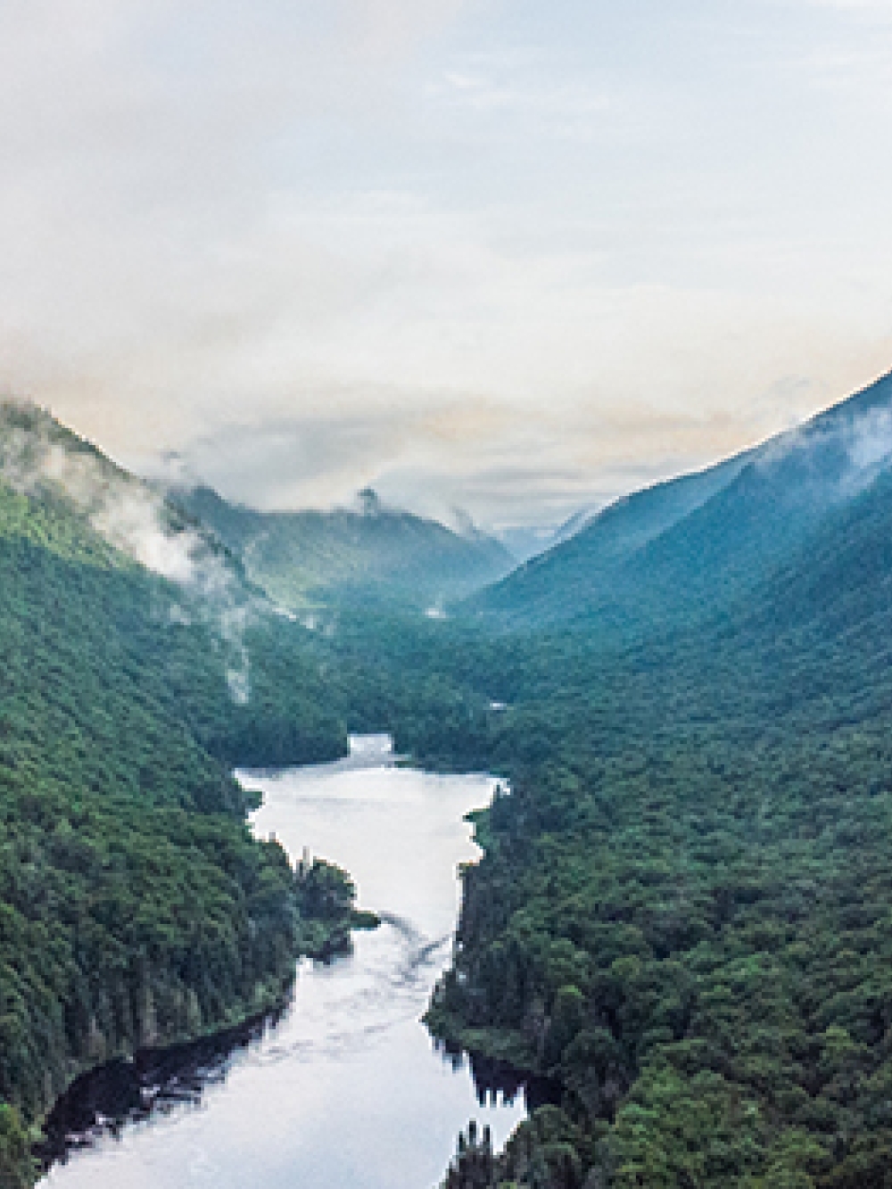 Vue aérienne d’une vallée verdoyante avec une rivière sinueuse entourée de montagnes boisées et de nuages bas.