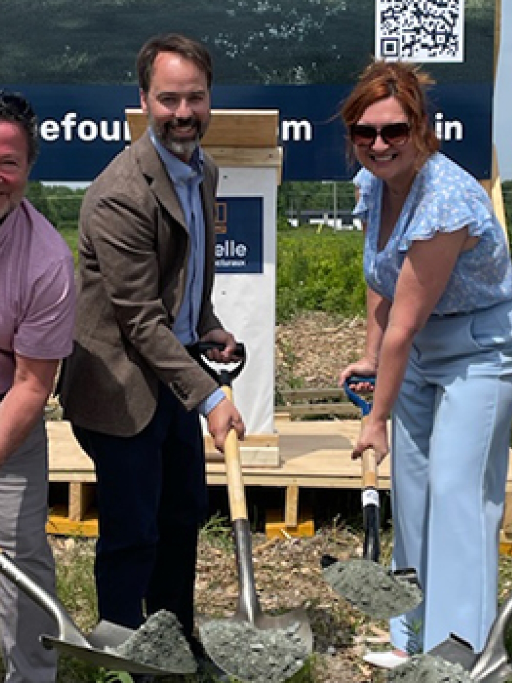 Quatre personnes en tenue professionnelle participent à une cérémonie de première pelletée de terre sur un terrain, tenant des pelles avec du gravier, devant une pancarte affichant le nom Fournelle.