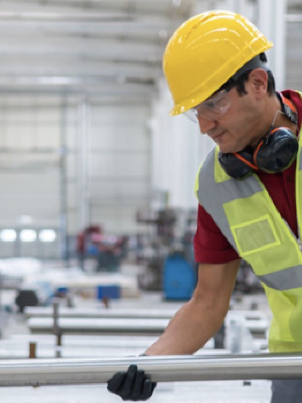 Travailleur portant un casque de sécurité et un gilet haute visibilité manipulant un long tube métallique dans un grand atelier industriel lumineux.