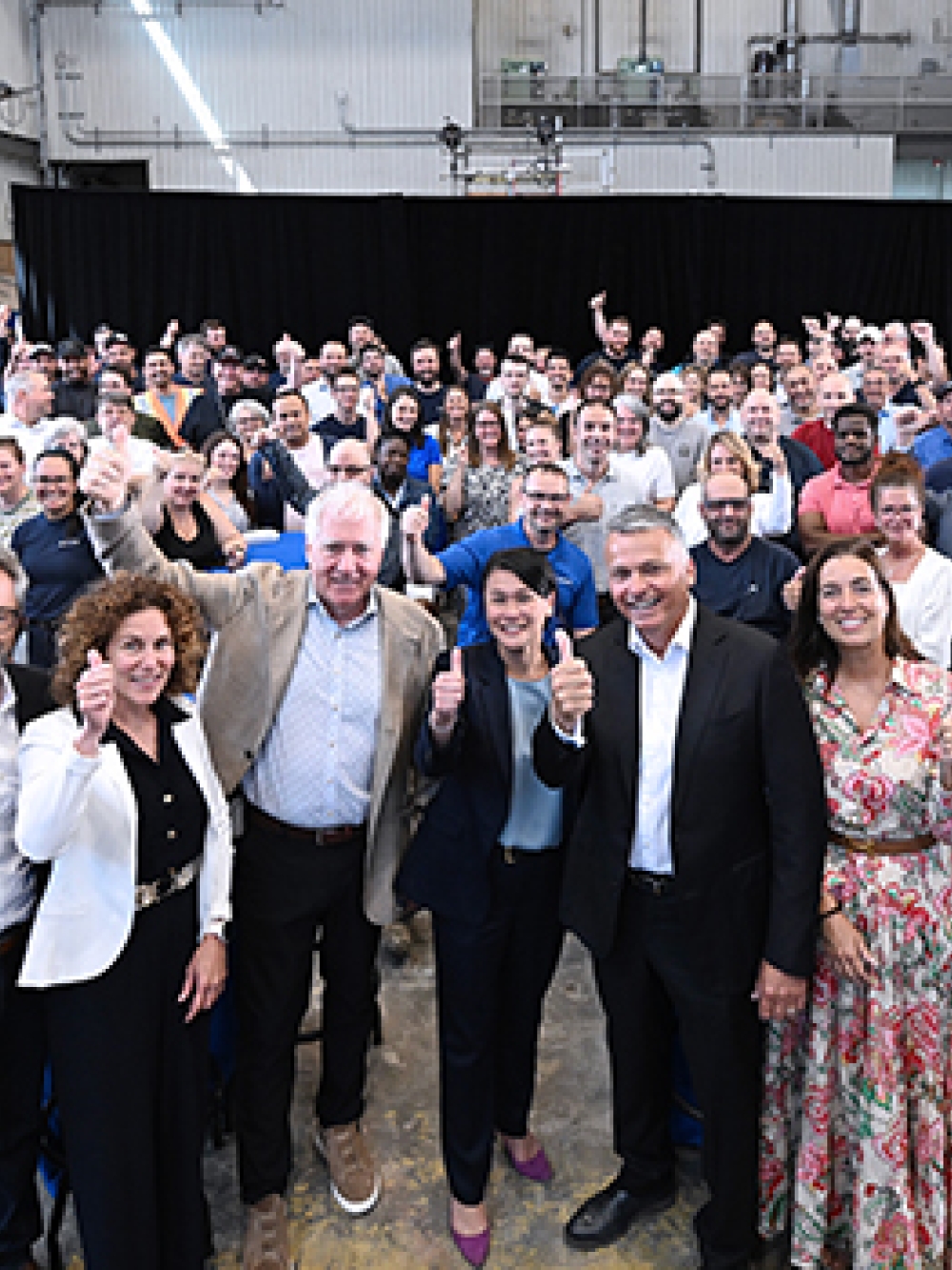 Grande assemblée de personnes réunies dans un vaste atelier industriel, certaines debout à l’avant et d’autres regroupées à l’arrière, avec des tables hautes recouvertes de nappes bleues et un rideau noir en fond de scène.