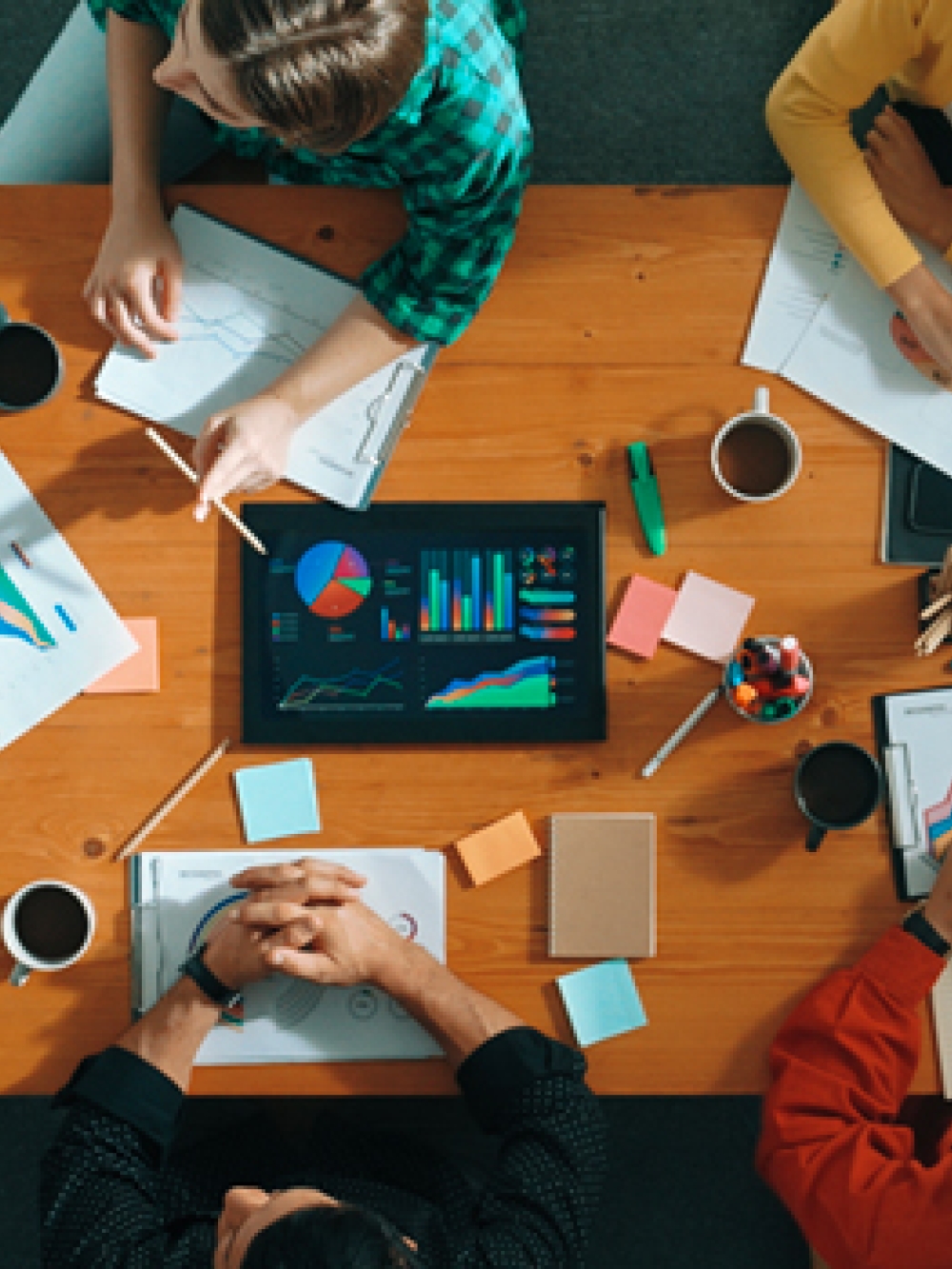 Vue aérienne d’un groupe de personnes autour d’une table de travail, échangeant sur des graphiques, documents et un écran affichant des données.
