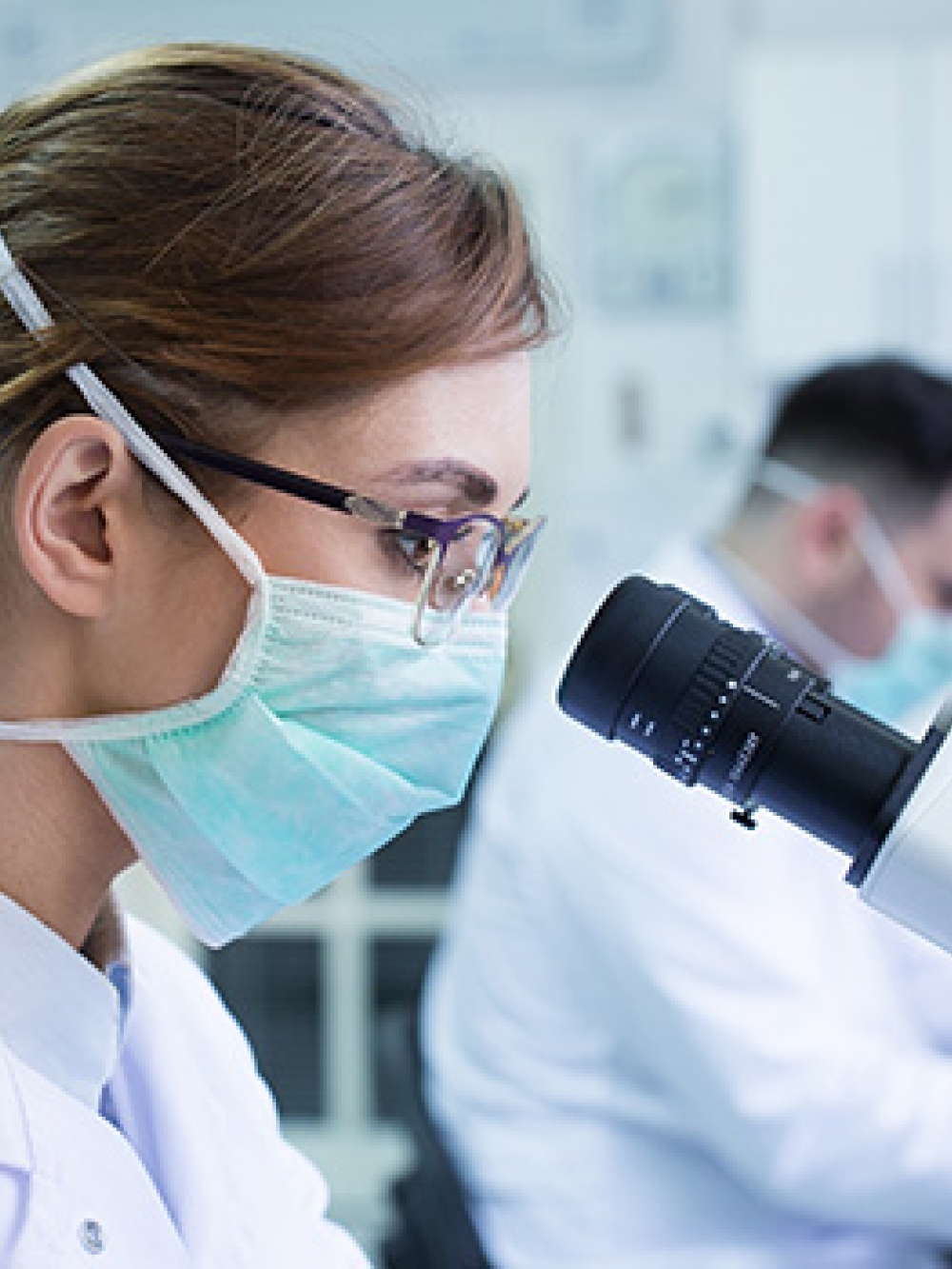 Personne en blouse de laboratoire observant un échantillon au microscope dans un environnement scientifique, avec un second technicien travaillant en arrière‑plan.