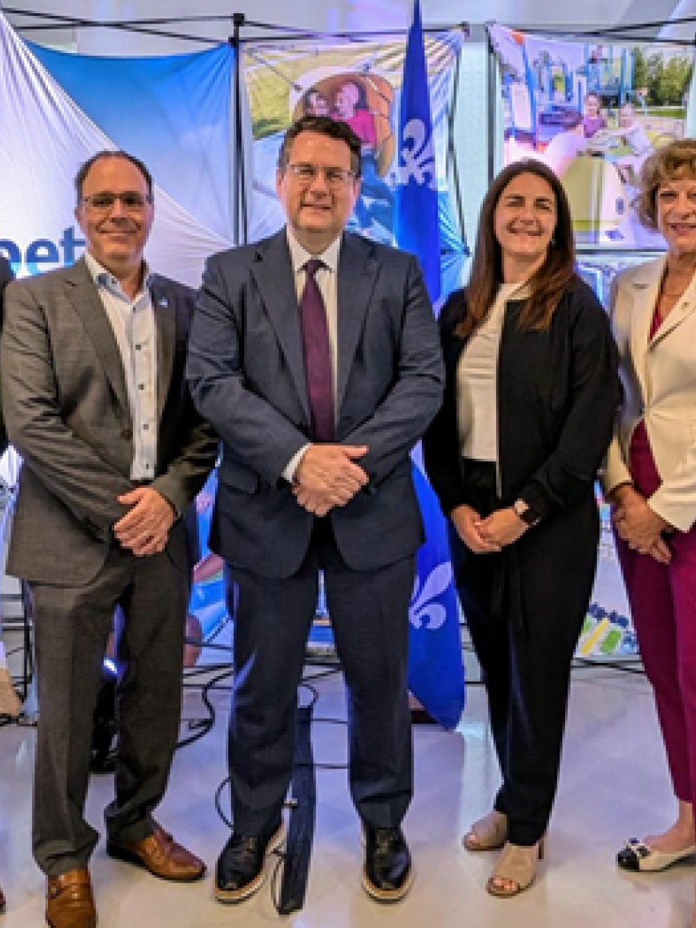 Groupe de six personnes debout devant un mur d’affiches illustrant des équipements de jeux extérieurs, avec un panneau portant le nom « Jambette » et des drapeaux du Québec en arrière-plan.