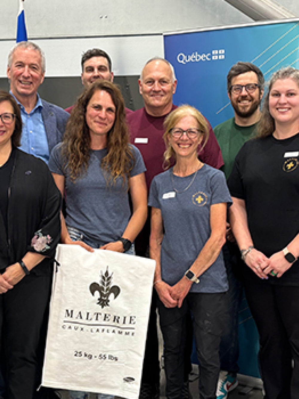 Groupe de personnes posant dans une installation industrielle, devant des équipements et deux panneaux dont un portant l’identité gouvernementale, avec au centre un sac de la Malterie Caux‑Laflamme.