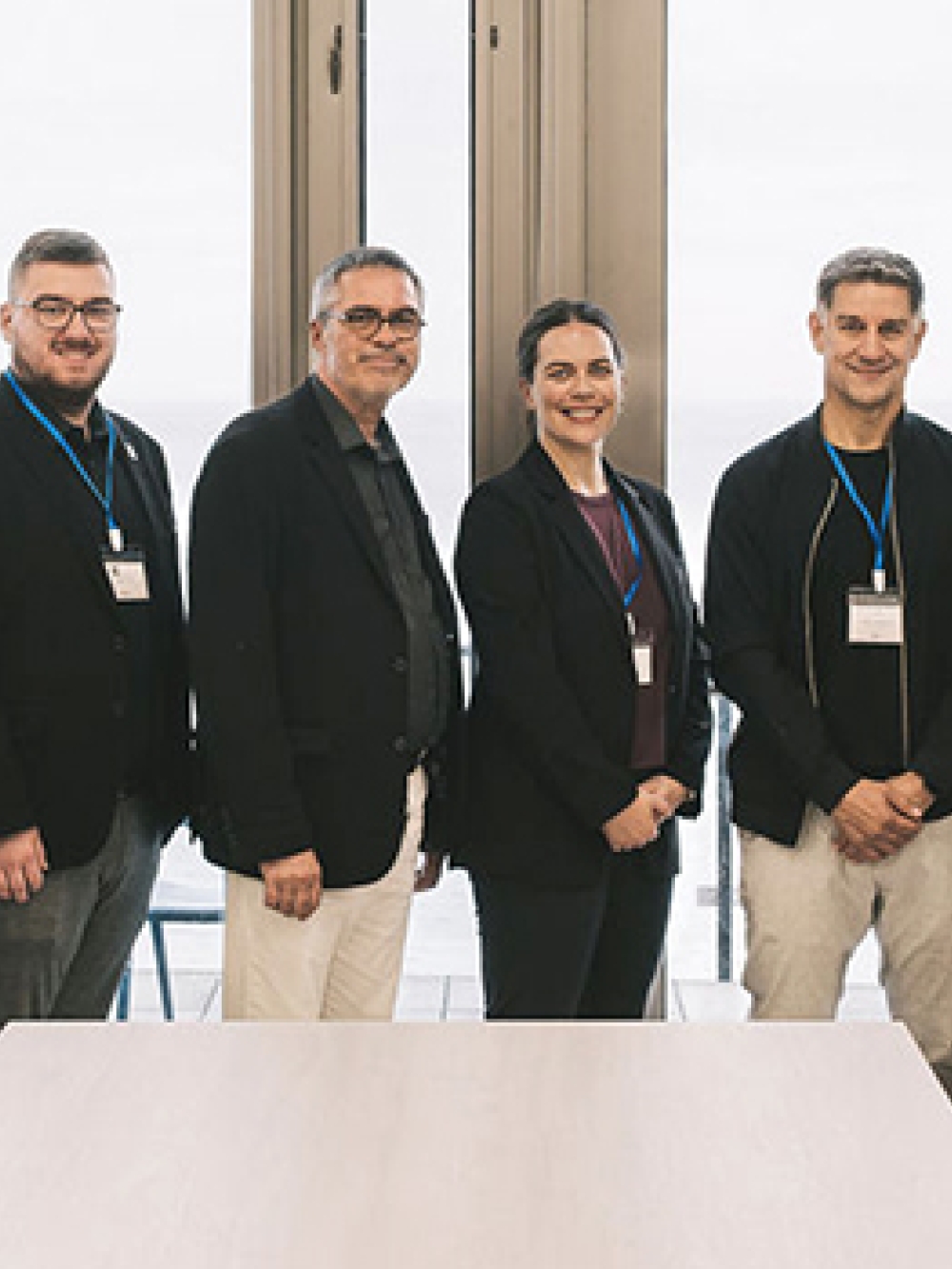 Groupe de personnes debout dans une salle lumineuse avec de grandes fenêtres donnant sur un paysage côtier, posant côte à côte devant une table.