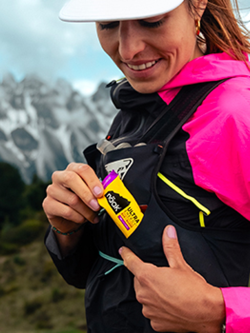Personne en tenue de sport en montagne insérant une barre énergétique Näak dans la poche d’un sac de course, avec des sommets enneigés en arrière-plan.