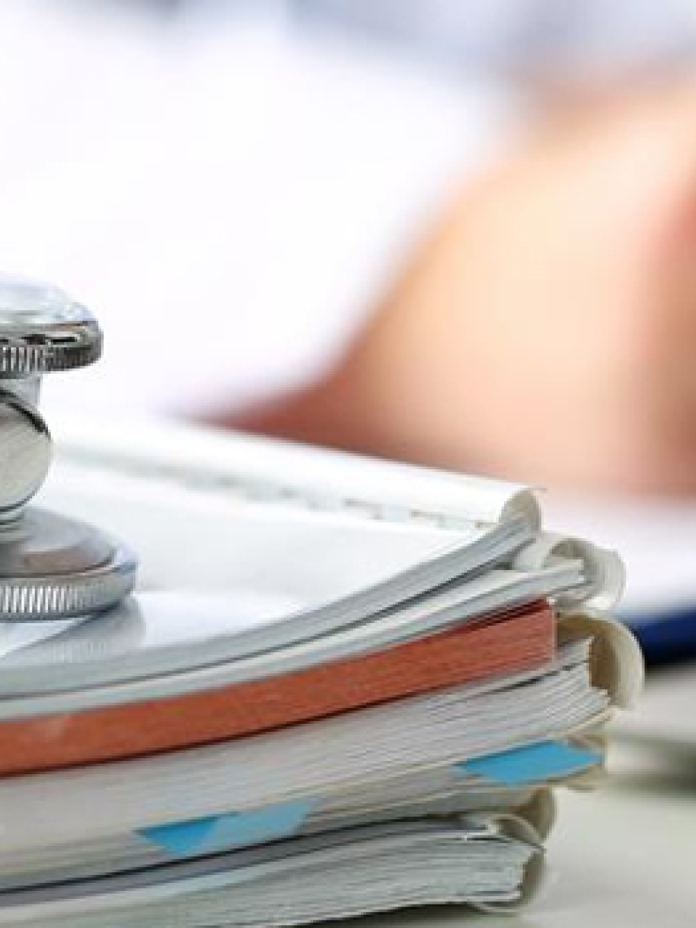 Stéthoscope posé sur une pile de dossiers médicaux, avec une personne écrivant à l’arrière‑plan.
