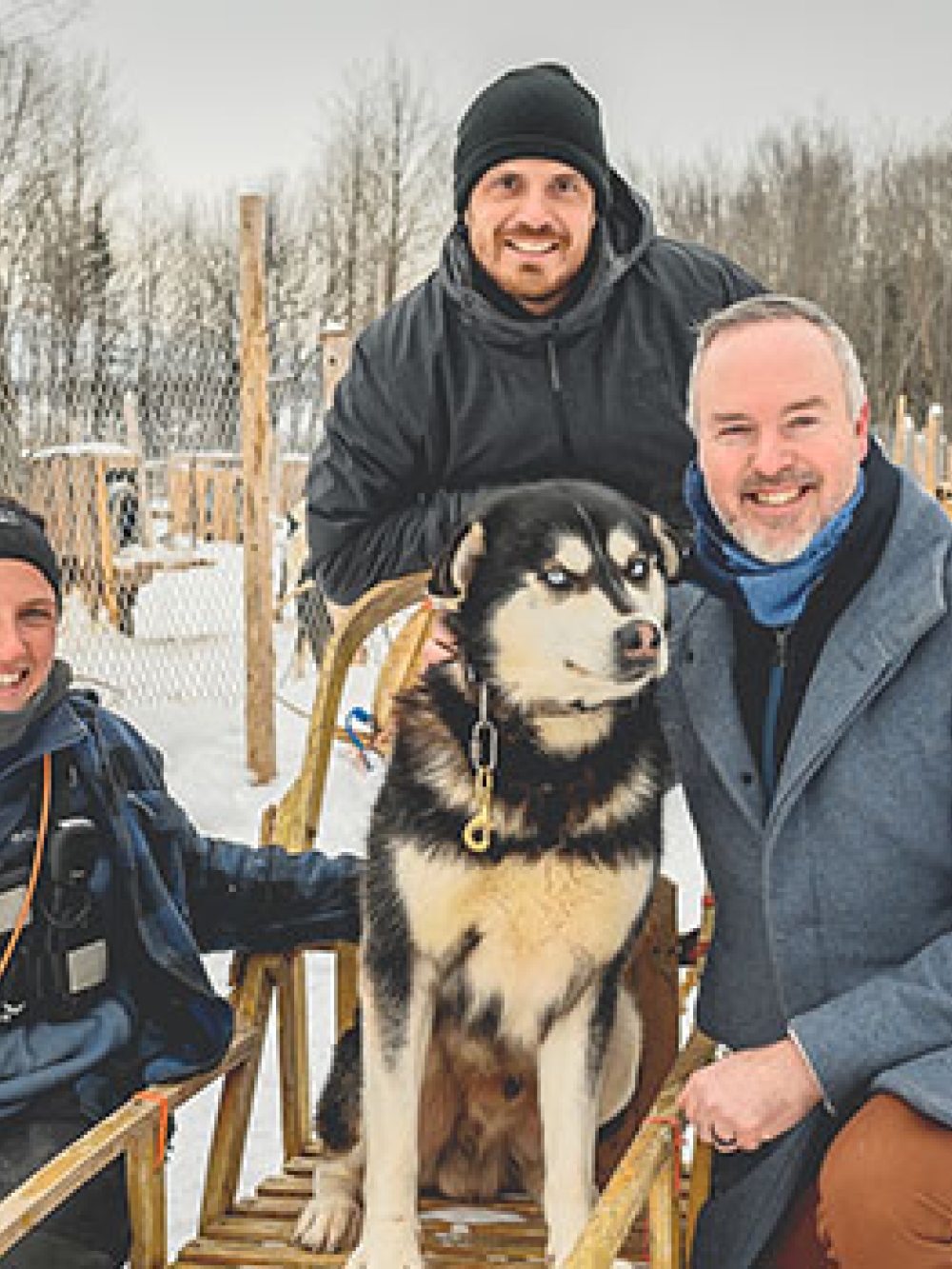 Trois personnes posant à l’extérieur dans un décor enneigé avec un grand chien de traîneau assis sur un traîneau, devant un chenil hivernal.