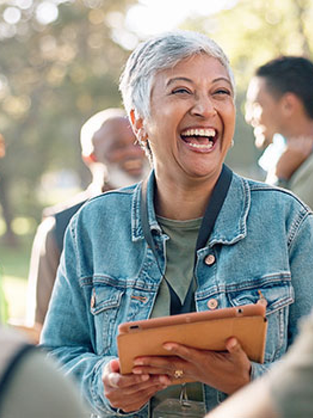 Personne tenant une tablette numérique et discutant avec un petit groupe en plein air, dans un environnement boisé et ensoleillé.