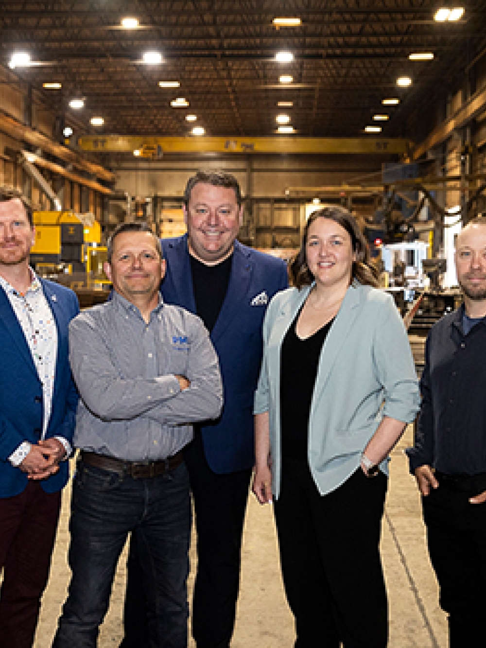 Groupe de personnes debout dans un grand atelier industriel, posant entre des équipements, des structures métalliques.