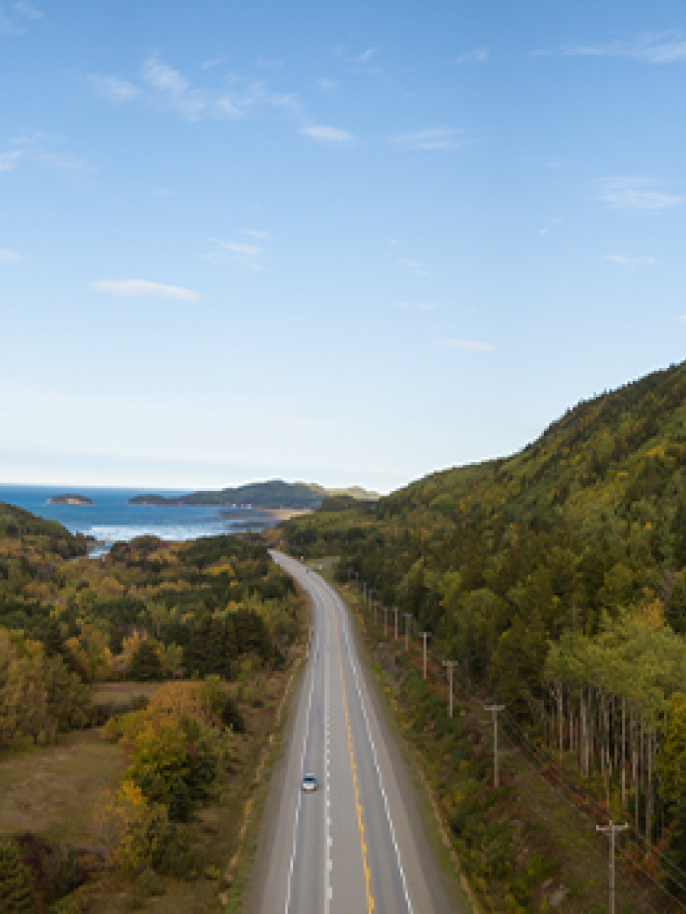 Route traversant une vallée entourée de collines boisées, avec la mer visible à l’horizon. 