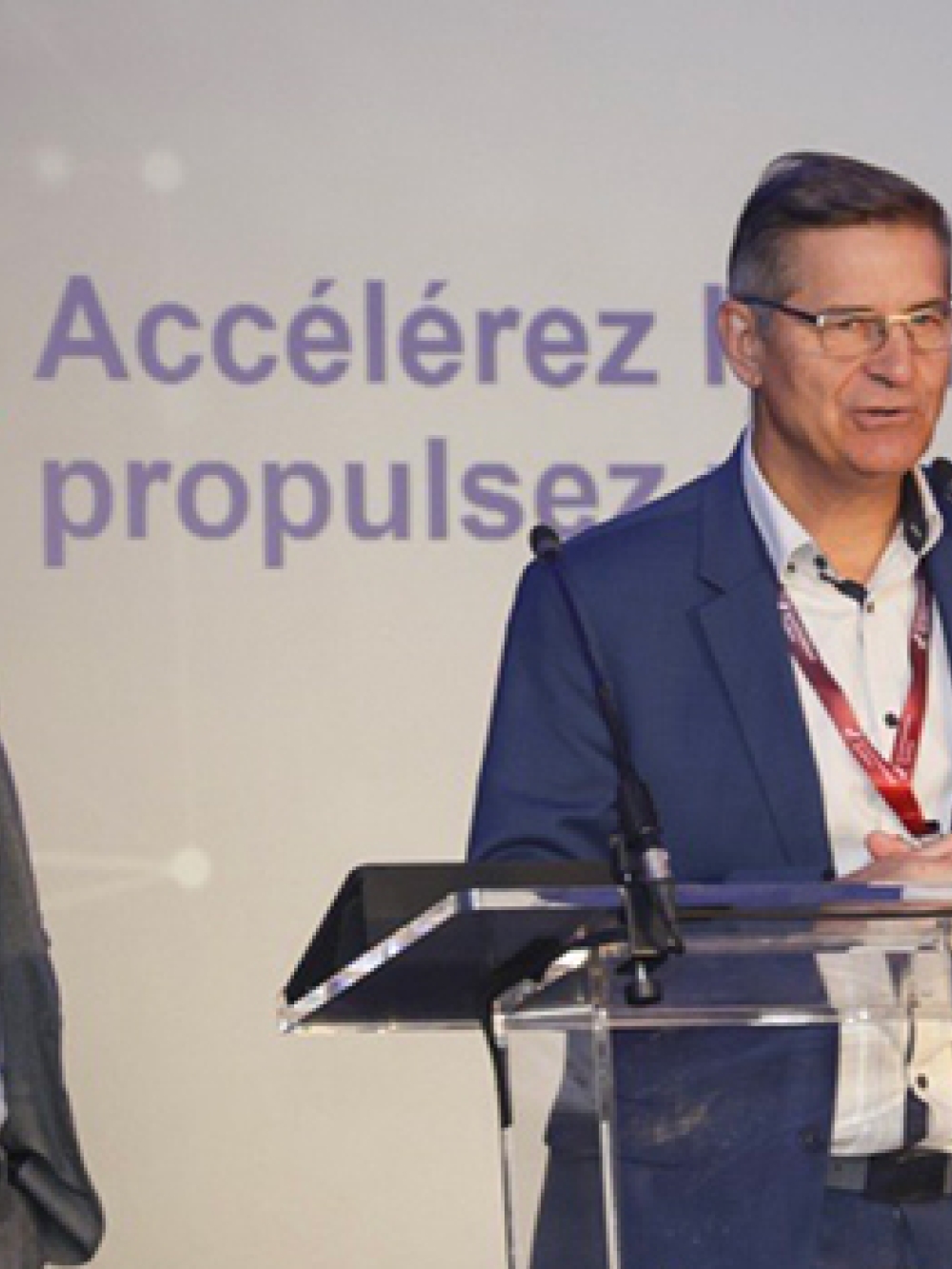Deux personnes en tenue professionnelle se tenant sur une scène, l’une derrière un lutrin, devant un écran affichant un message sur l’innovation et la productivité. 