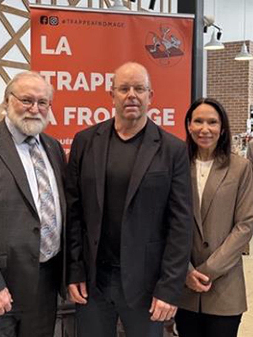 Groupe de cinq personnes posant dans un commerce de fromagerie, devant une affiche « La Trappe à Fromage » et un drapeau du Québec, avec des étagères de produits à droite.