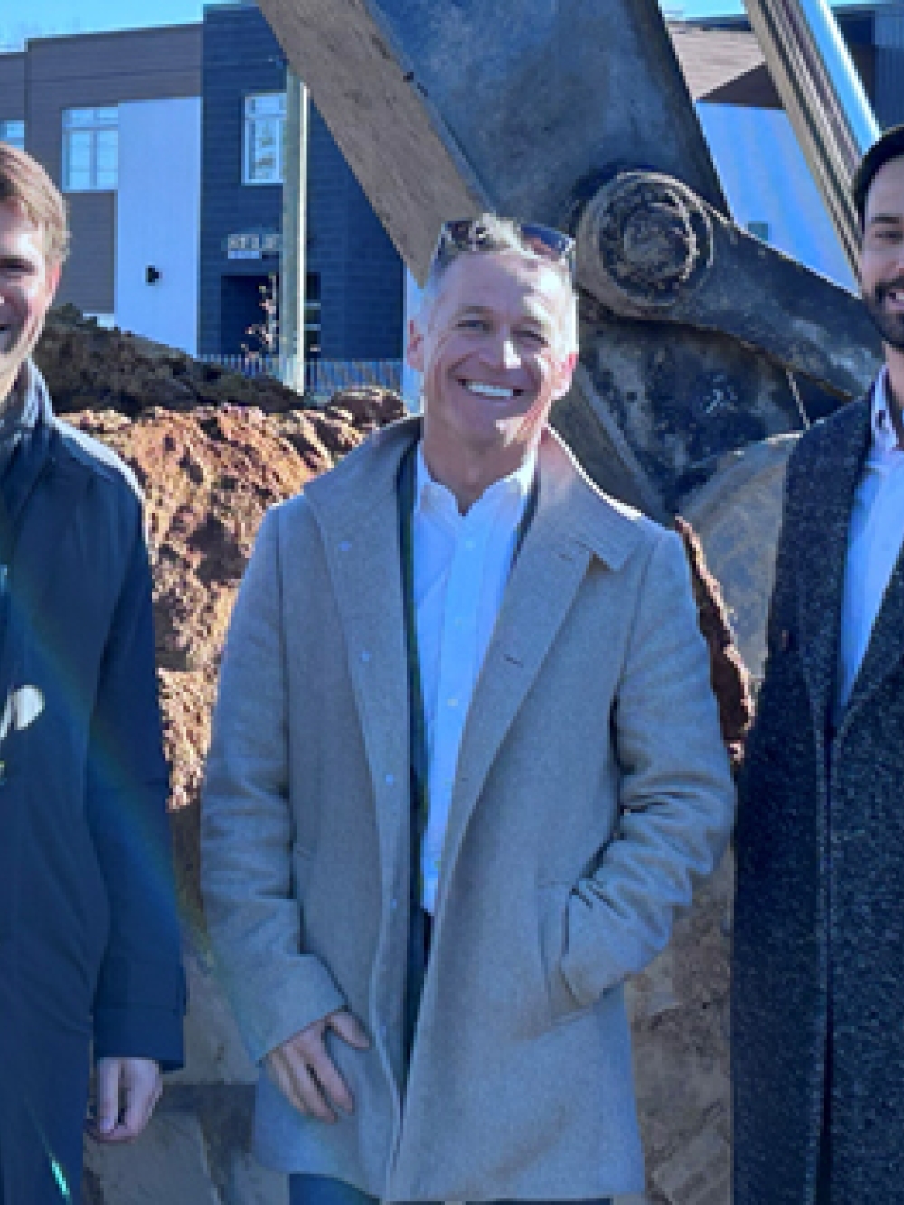 Groupe de personnes debout sur un chantier devant une pelle mécanique et un tas de terre, avec des bâtiments industriels en arrière-plan.