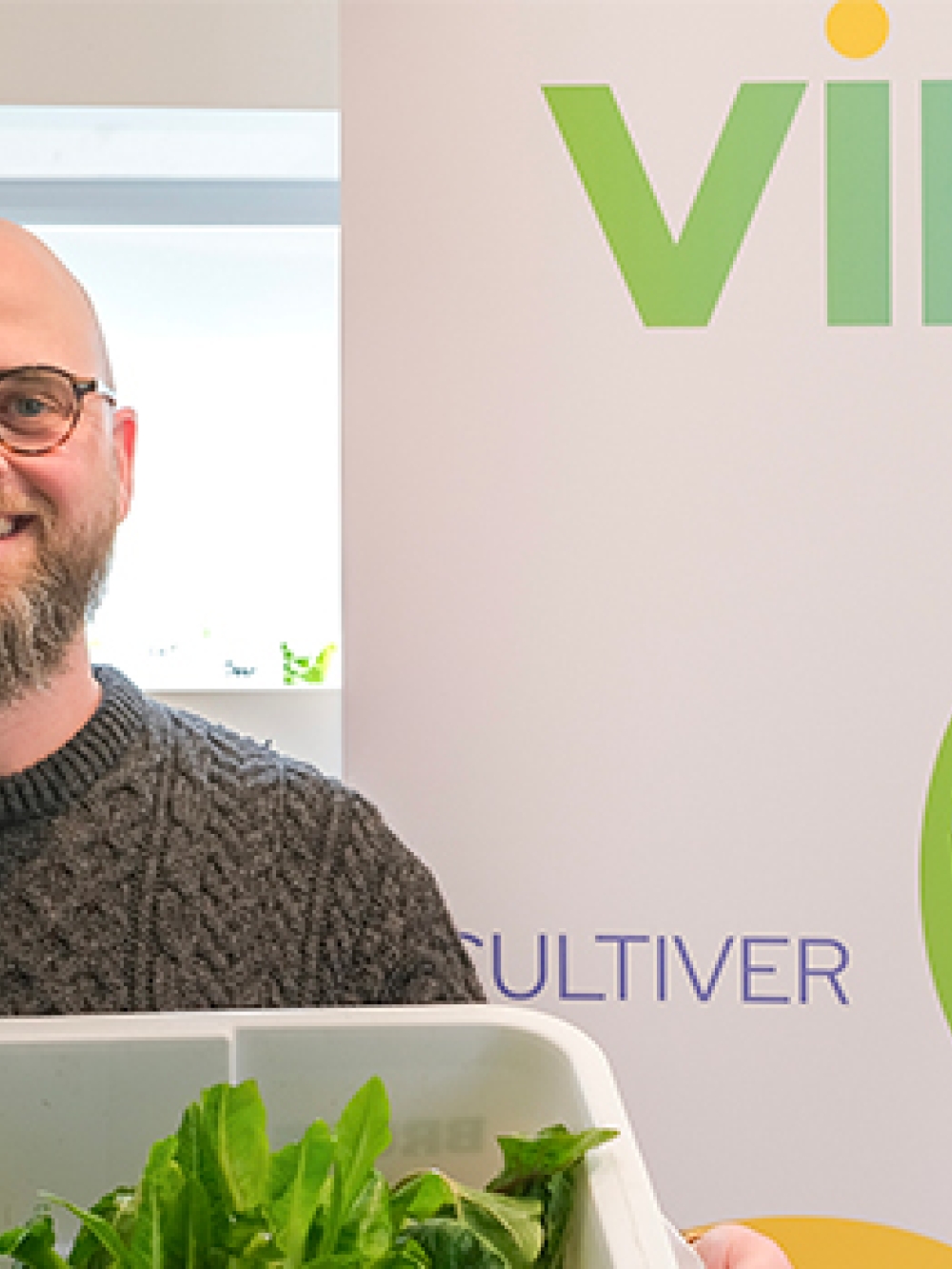Personne tenant un bac de légumes frais devant une bannière de Vireo, dans un environnement d’agriculture urbaine hydroponique.