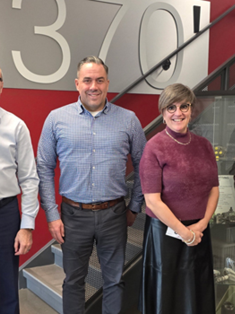 Groupe de personnes posant dans un espace industriel moderne, devant un mur rouge portant l’inscription « 370! », près d’un escalier métallique et de vitrines présentant des pièces mécaniques.