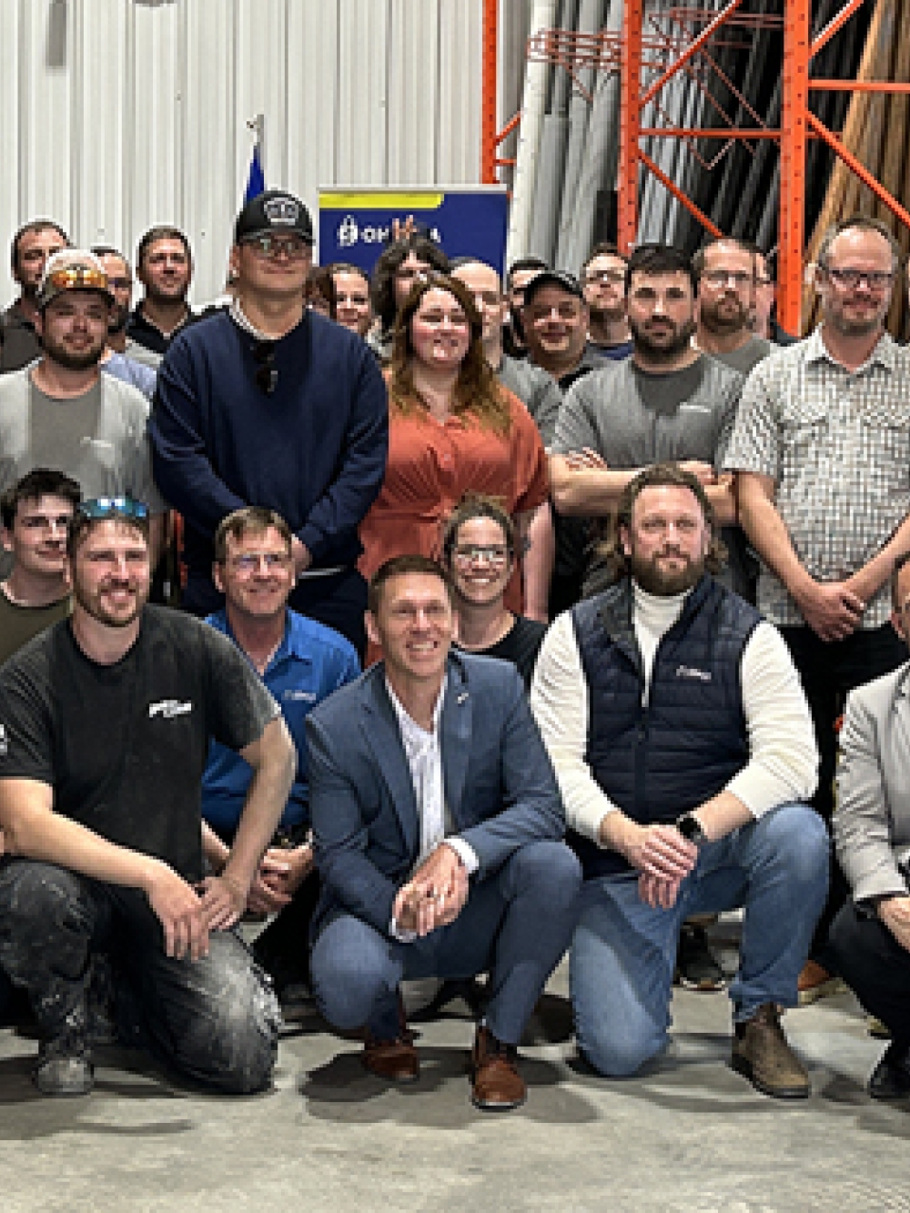 Grand groupe de personnes posant pour une photo à l’intérieur d’un entrepôt industriel, entouré de rayonnages métalliques orange et d’équipements de manutention.