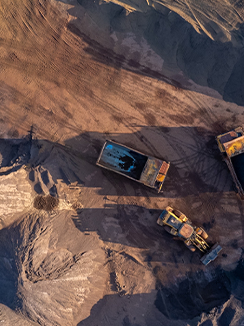 Vue aérienne d’un site minier où plusieurs camions et machineries lourdes déplacent et chargent des matériaux sur un terrain de sable et de gravier. 