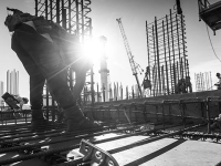 Travailleurs sur un chantier assemblant des barres d’armature en acier, avec le soleil en arrière‑plan et des structures verticales en construction.