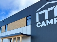 Façade moderne d’un bâtiment industriel gris et bois portant le logo « NT CAMPS », sous un ciel bleu.
