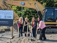 Quatre personnes tenant des pelles lors d’une cérémonie de première pelletée devant un panneau EcoloPharm et une pelle mécanique CAT sur un chantier.