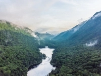 Vue aérienne d’une vallée entourée de montagnes couvertes de forêt dense, avec une rivière sinuant au centre sous une brume matinale. 
