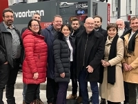 Groupe de personnes debout dans un atelier industriel devant une grande machine de production, portant des manteaux d’hiver et des vêtements de travail.