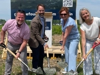 Quatre personnes en tenue professionnelle participent à une cérémonie de première pelletée de terre sur un terrain, tenant des pelles avec du gravier, devant une pancarte affichant le nom Fournelle.