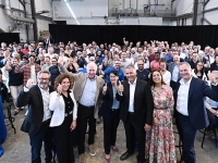 Grande assemblée de personnes réunies dans un vaste atelier industriel, certaines debout à l’avant et d’autres regroupées à l’arrière, avec des tables hautes recouvertes de nappes bleues et un rideau noir en fond de scène.