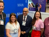 Cinq personnes debout à l’intérieur lors d’un événement professionnel, portant des badges autour du cou, devant un panneau bleu avec le mot « MAIN ».