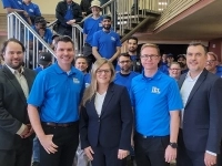 Groupe de personnes réunies dans un hall intérieur, plusieurs portant des chandails bleus avec le logo IEL, posant devant un escalier métallique dans un environnement industriel.