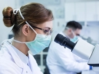 Personne en blouse de laboratoire observant un échantillon au microscope dans un environnement scientifique, avec un second technicien travaillant en arrière‑plan.