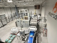 Vue d’ensemble d’une installation industrielle automatisée avec convoyeurs, postes d’emballage, équipements de manutention et sacs de produits, dans un entrepôt portant le logo Intersand sur le mur.