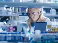 Personne en laboratoire travaillant avec un microscope, entourée de matériel scientifique et de flacons.