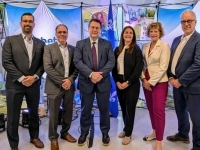 Groupe de six personnes debout devant un mur d’affiches illustrant des équipements de jeux extérieurs, avec un panneau portant le nom « Jambette » et des drapeaux du Québec en arrière-plan.