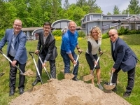 Groupe de personnes participant à une pelletée de terre symbolique sur un terrain gazonné, chacun tenant une pelle remplie de sable, avec des bâtiments et des arbres en arrière‑plan.