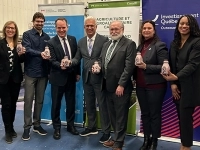 Groupe de sept personnes posant dans une usine devant des bannières gouvernementales, chacune tenant une petite bouteille blanche, avec des drapeaux du Québec et du Canada de chaque côté.