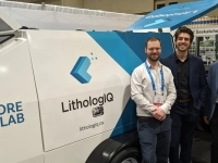 Unité mobile blanche avec le logo LithologIQ et l’inscription « Mobile Core Scanner Lab », exposée dans un salon professionnel, avec trois personnes debout à côté et des kiosques en arrière-plan.
