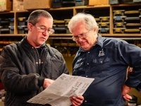 Deux personnes dans un atelier mécanique discutant et examinant un document technique, entourées d’étagères remplies de boîtes et d’outils.