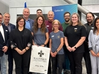 Groupe de personnes posant dans une installation industrielle, devant des équipements et deux panneaux dont un portant l’identité gouvernementale, avec au centre un sac de la Malterie Caux‑Laflamme.